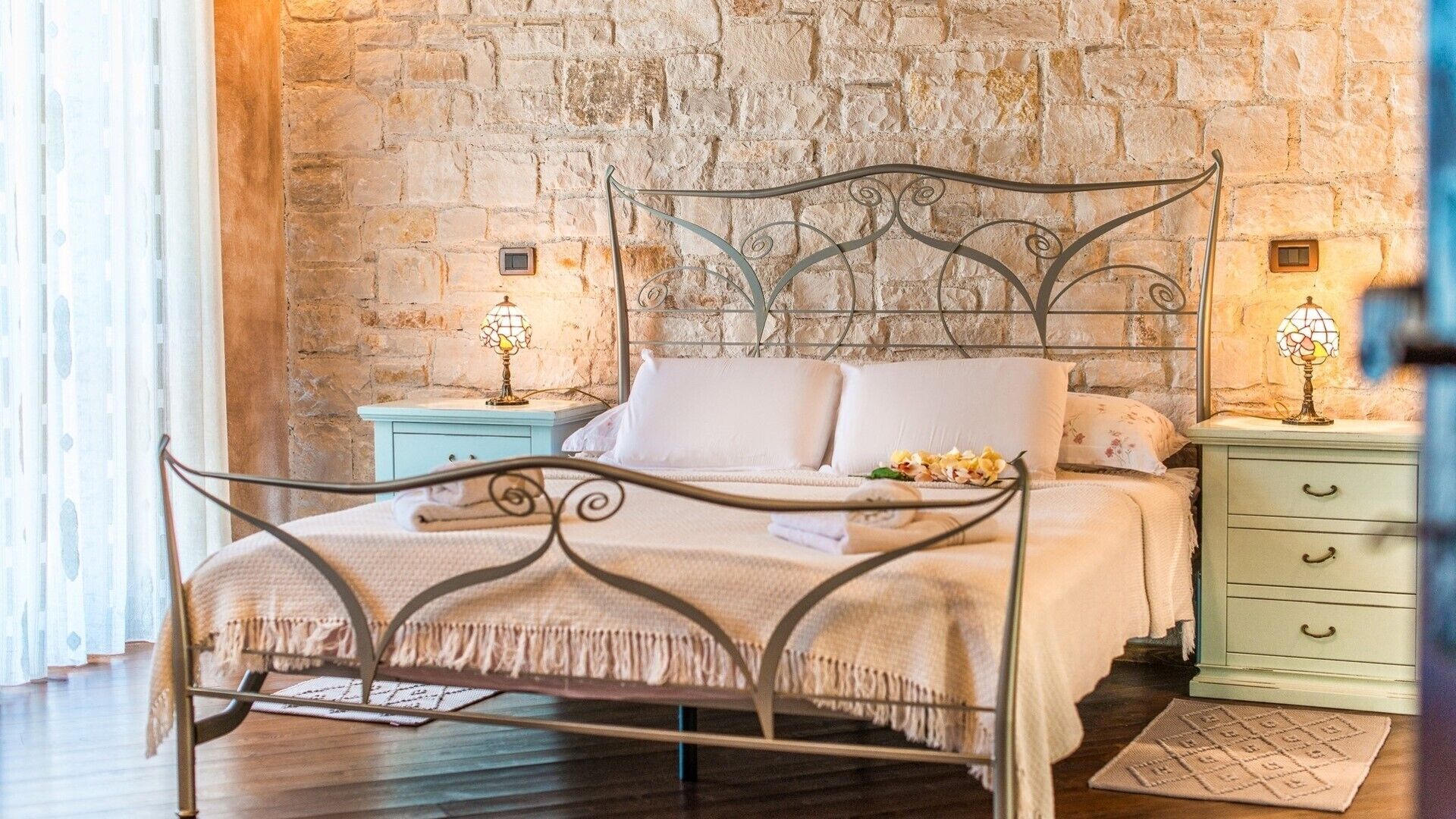 A bed with a metal frame, a headboard, and a footboard, set against a stone wall. There are two bedside tables with lamps, a window with curtains, and a wooden floor.