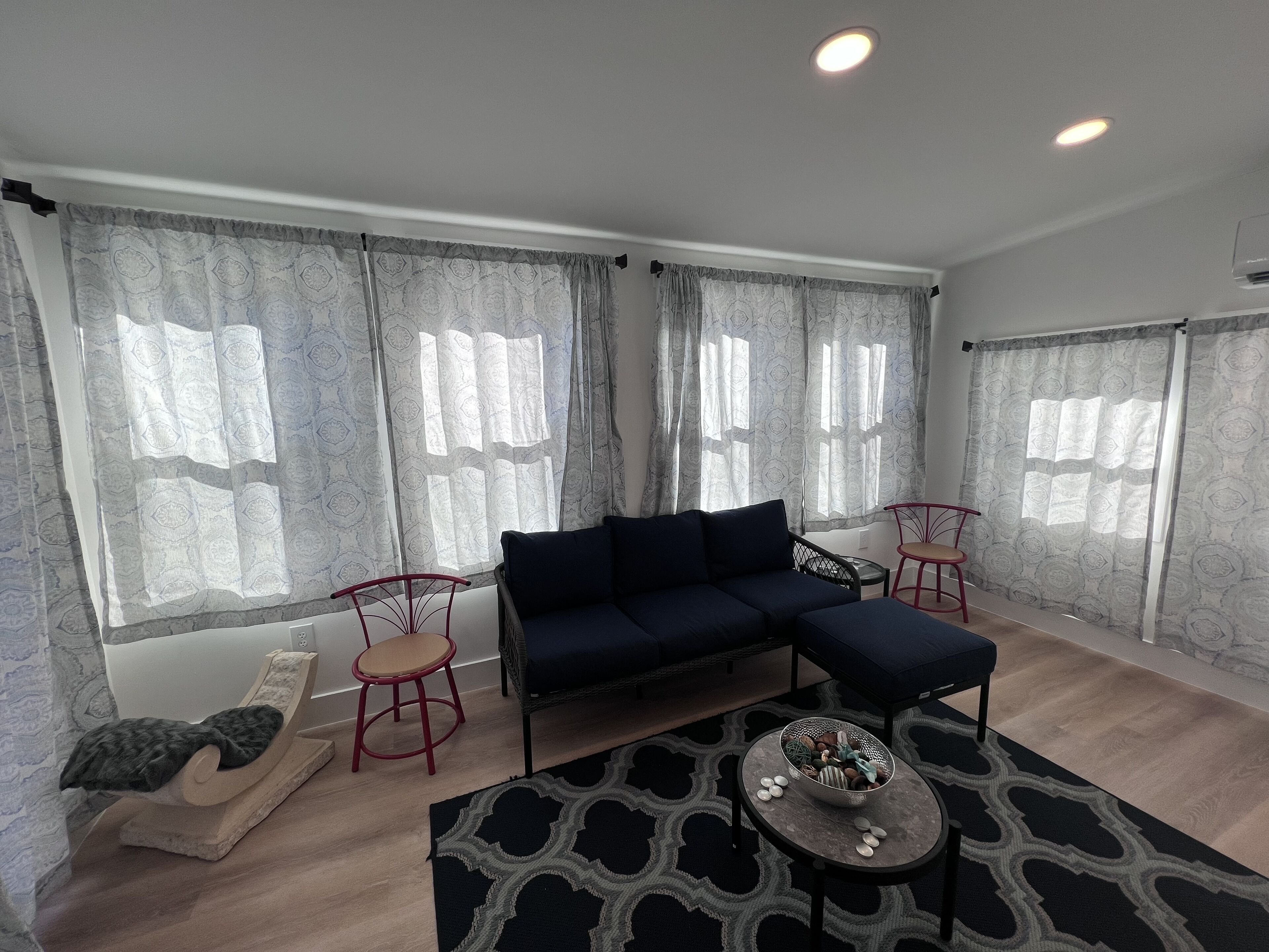 Un salon moderne avec un canapé bleu foncé, deux fauteuils rouges, une petite table ronde sur laquelle se trouve un bol de collations et un tapis à motifs.