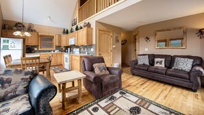 A spacious living area with a high ceiling, a wooden staircase, a dining table with chairs, a leather sofa, and a large window.