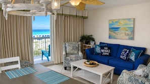 A living room with a view of the ocean, a blue sofa with nautical-themed pillows, and a dining area with striped placemats.