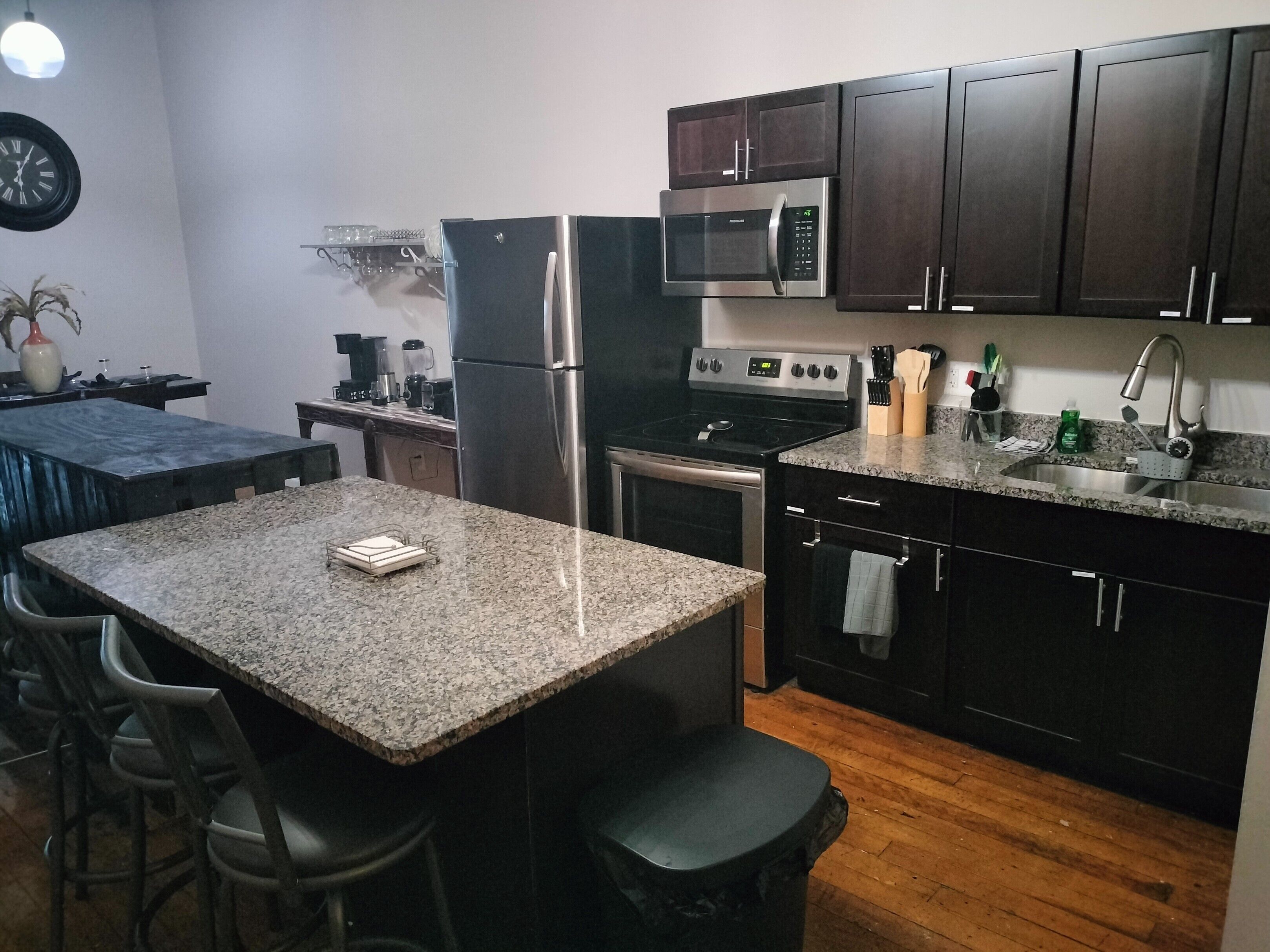 A compact kitchen with dark cabinets, a granite countertop, stainless steel appliances, and a small dining area with bar stools.