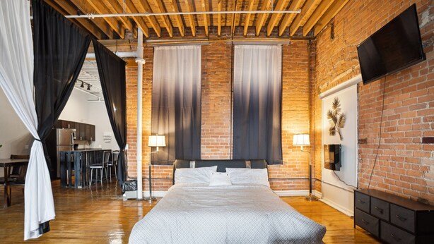 A modern bedroom with a large bed, a wooden ceiling, brick walls, and a flat-screen TV mounted on the wall. - Downtown Detroit Loft - Fully equipped & Absolutely Gorgeous theme by RedAwning (Detroit)