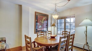 A dining area with a round wooden table surrounded by six wooden chairs, a hanging light fixture, a framed poster on the wall, and a window with a view of buildings outside. - 1BR Plus Den, Heart of Beaver Creek Village 1 Bedroom Condo by RedAwning (Avon)