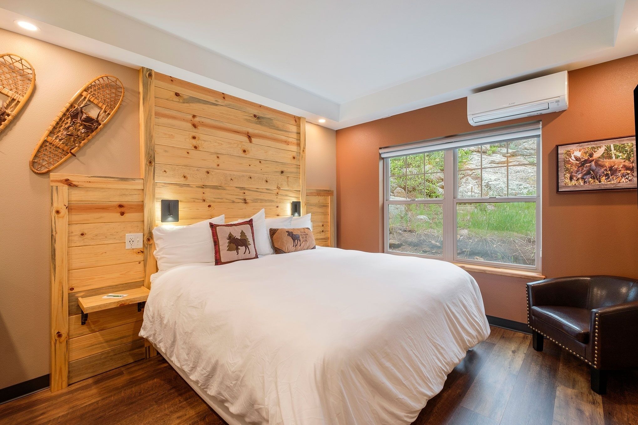 A bedroom with a wooden headboard, a large bed with white bedding, a window with a view of greenery, a brown leather chair, and an air conditioning unit.