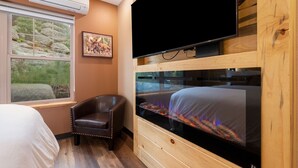 A bedroom with a bed, a TV, a fireplace, a chair, and a window with a view of rocks and greenery.