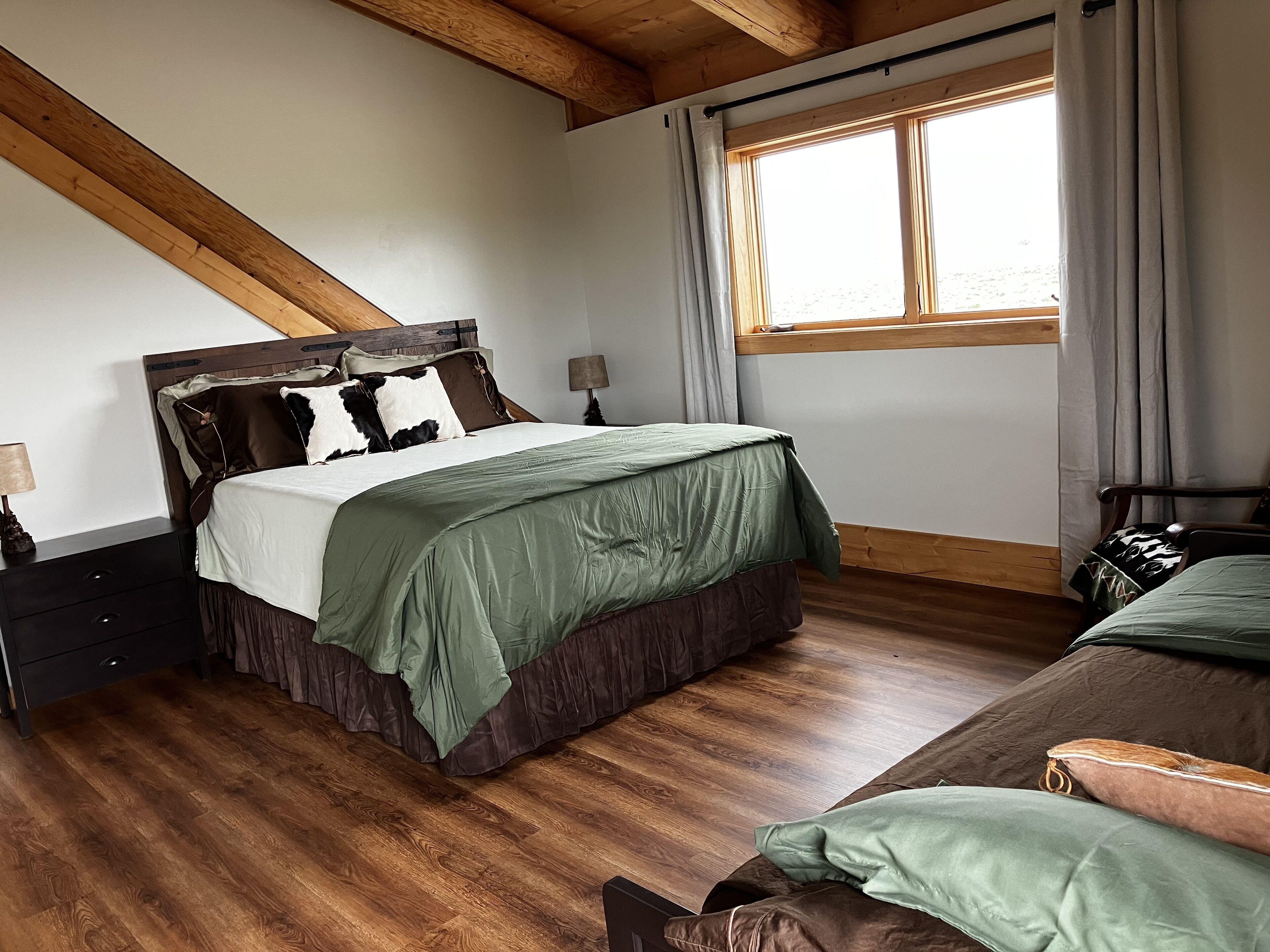 A bedroom with a wooden ceiling, a large bed with a green bedspread, a nightstand with a lamp, and a window with curtains.