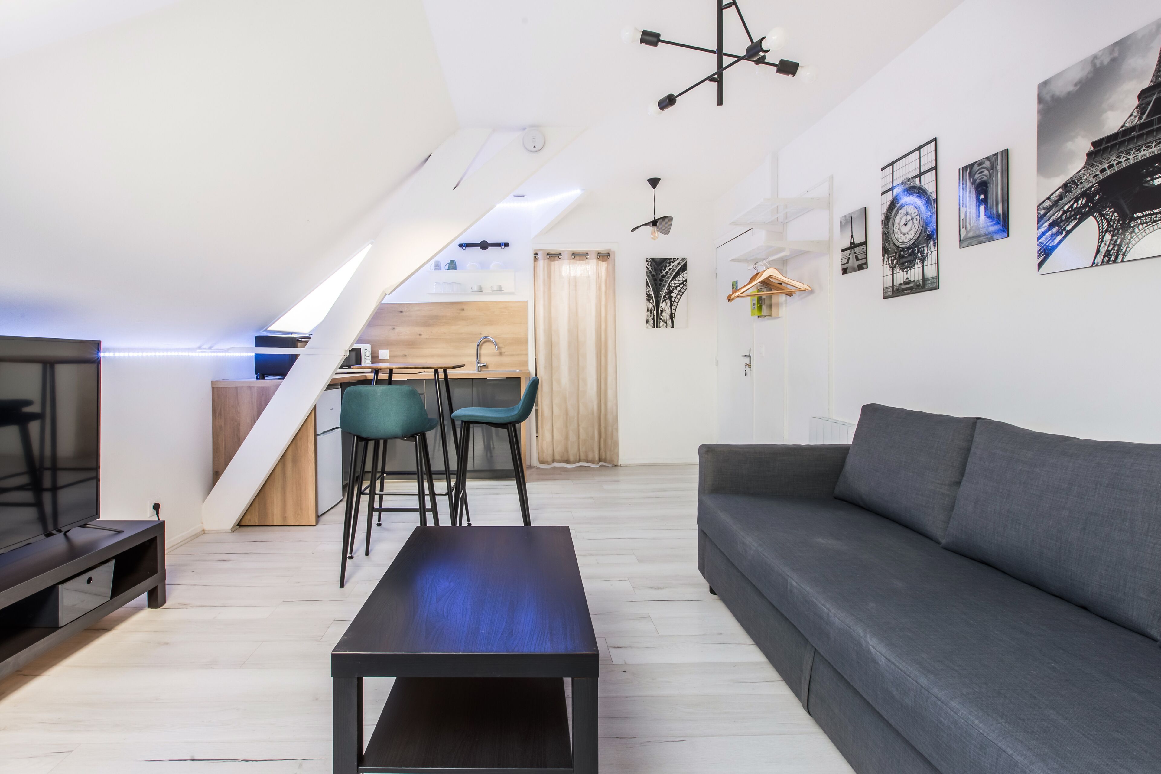 Ein modernes Wohnzimmer mit einer Couch, einem Couchtisch und einem Fernseher. Im Hintergrund ist eine Küchenzeile mit einem Esstisch und Stühlen zu sehen.