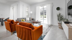 Ein modernes Wohnzimmer mit einem grauen Sofa, orangefarbenen Sesseln, einem Couchtisch und einem großen Fenster mit weißen Vorhängen.