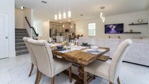 A dining area with a wooden table set for four, surrounded by upholstered chairs. A kitchen is visible in the background, and a flat-screen TV is mounted on the wall.