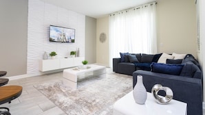 Una sala de estar moderna con un mueble blanco para entretenimiento, un sofá gris y una mesa de centro de vidrio.