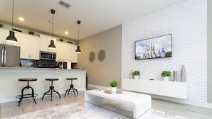 Cocina moderna con armarios blancos, un refrigerador de acero inoxidable y un microondas. Hay tres taburetes de bar y una mesa de centro blanca con una planta. Un televisor de pantalla plana está montado en una pared con textura.
