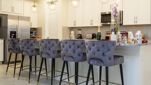 A modern kitchen with white cabinets, a stainless steel refrigerator, a microwave, and a bar area with tufted bar stools. - Last Minute Disney! Private Pool, Hot Tub, Game Room & Themed Bedrooms. Book Now! #90507 (Orlando)