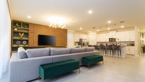A modern living room with a large sofa, green ottomans, a flat-screen TV, wooden shelving, and a kitchen area with bar stools. - Last Minute Disney! Private Pool, Hot Tub, Game Room & Themed Bedrooms. Book Now! #90507 (Orlando)