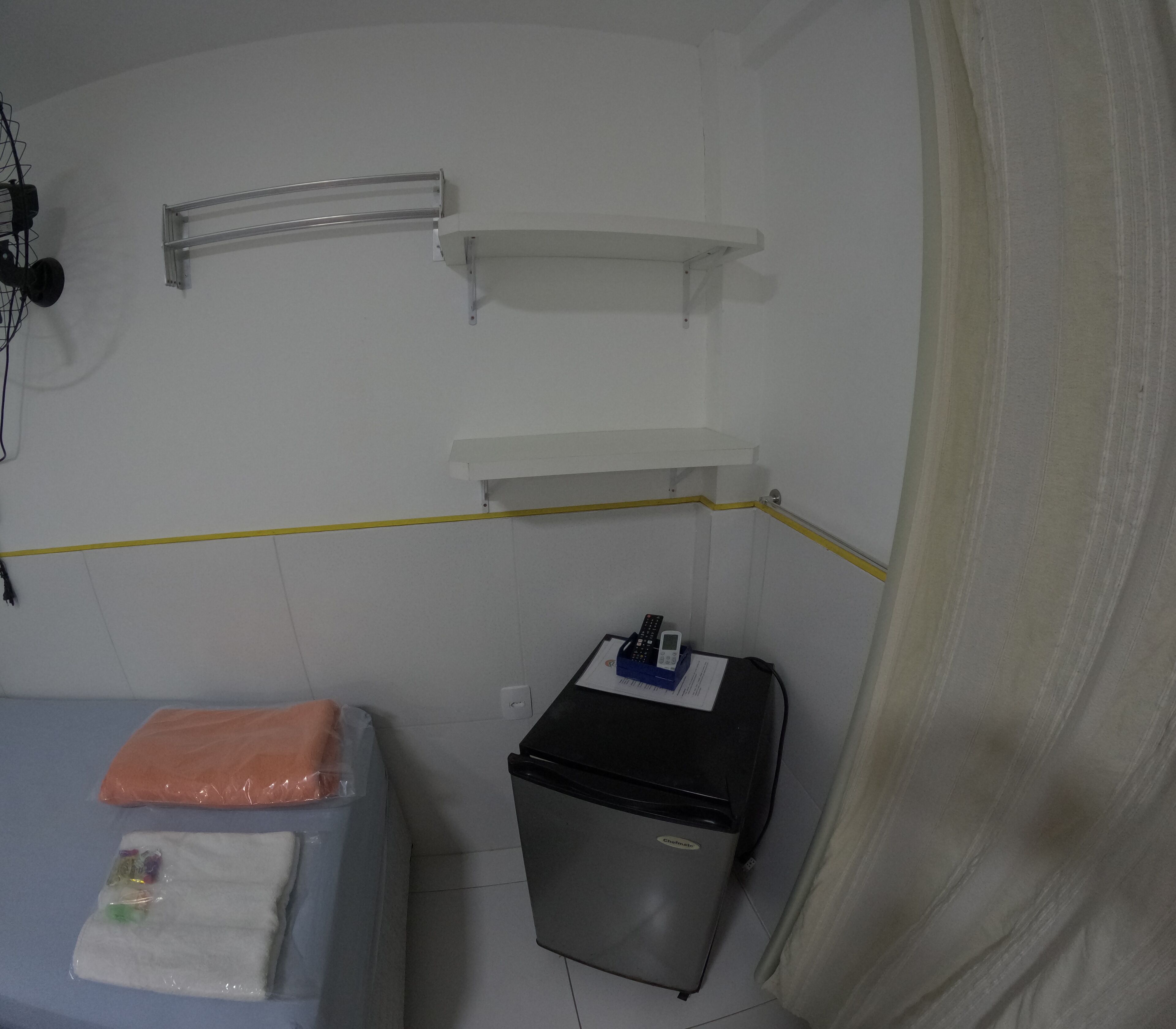 A small, sparsely furnished room with a bed, a trash bin, a heater, and a shelf unit.