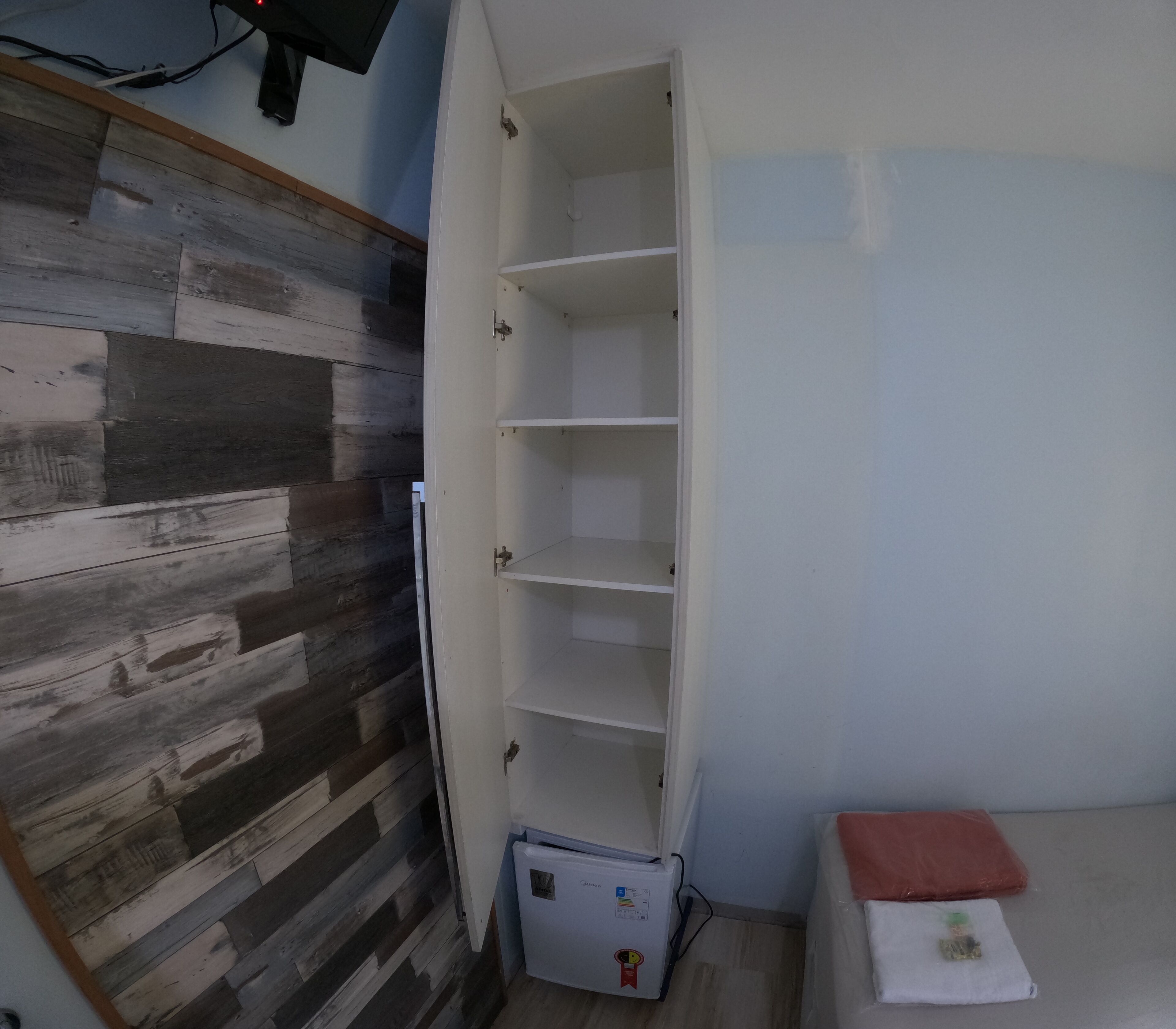 A room with a wooden wall, a white cabinet with multiple shelves, a small refrigerator, and a red cushion on the floor.