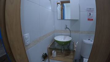 A bathroom with a wooden vanity, a green sink, a mirror, a toilet, and a trash bin.