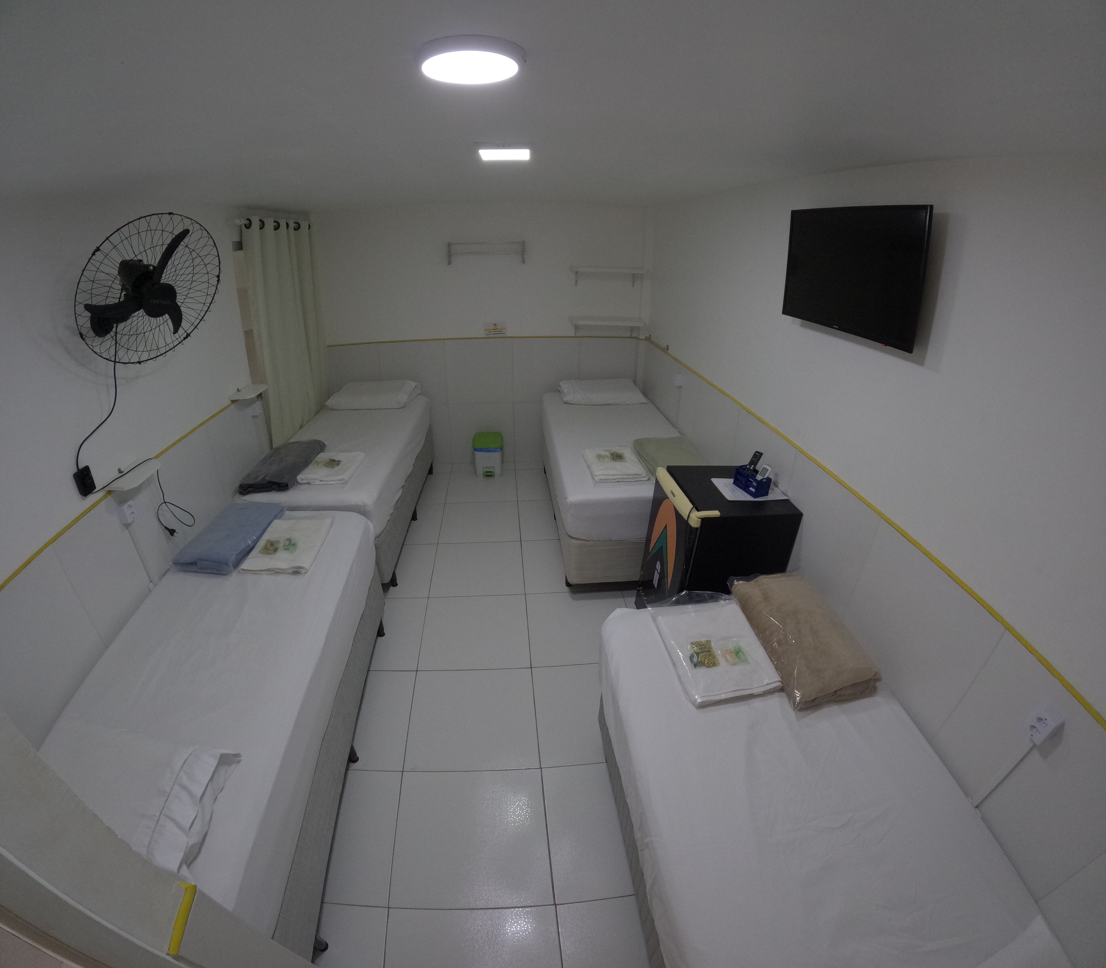 A room with three single beds, a wall-mounted TV, a ceiling fan, and a small trash bin.