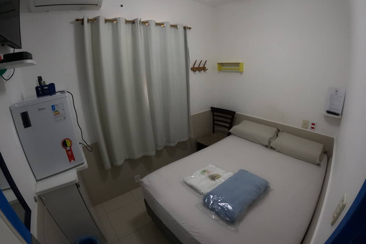 Una habitación de hotel con cama, ventilador de techo, equipo de aire acondicionado, una mesita, una silla, un cubo azul y un estante en la pared.