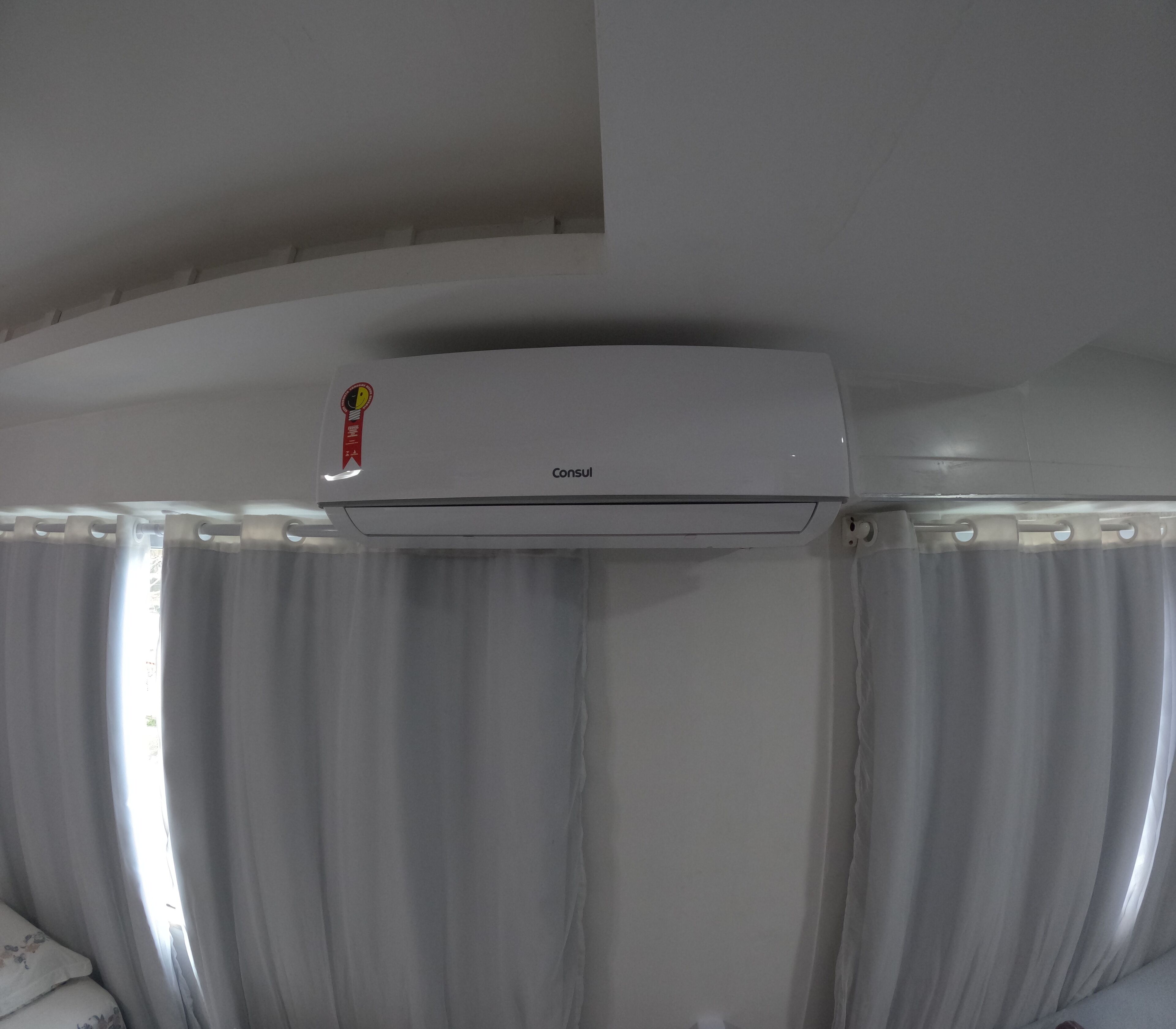 A white air conditioning unit mounted on a ceiling above a window with white curtains.