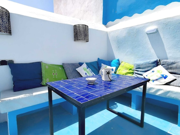 Un espace extérieur ensoleillé, aménagé avec des sièges, une table aux carreaux bleus, des coussins et une fresque murale.