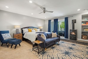 A modern bedroom with a bed, a sofa, a chair, a small table, a fireplace, and a view of the outdoors.