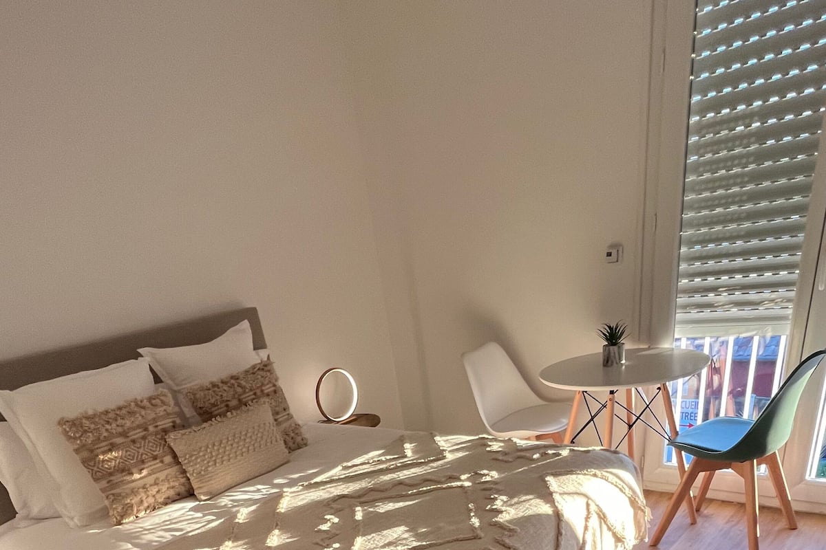A bedroom with a large bed, a small table with a chair, and a window with blinds.