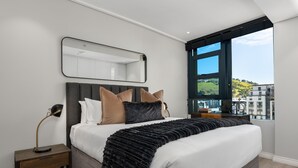 Ein modernes Schlafzimmer mit einem großen Bett, einem Nachttisch, einem Spiegel und Blick auf Gebäude durch ein großes Fenster.