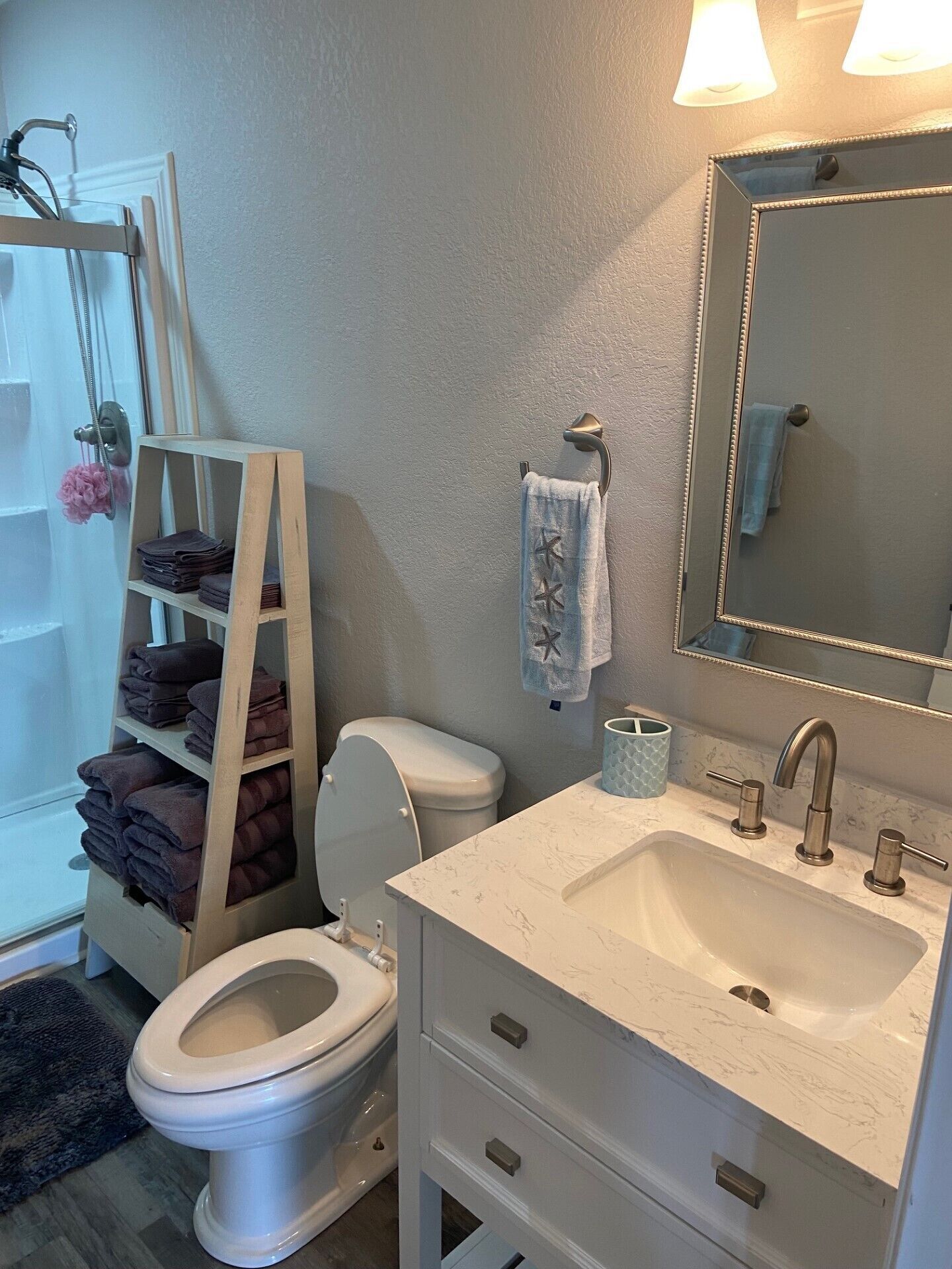 Une salle de bain avec des toilettes blanches, un meuble-lavabo avec un évier et un miroir, un porte-serviettes et une étagère avec des serviettes pliées.