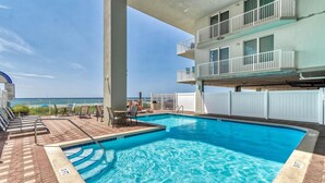 A swimming pool with a view of the ocean, a covered seating area, and a multi-story building with balconies.