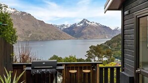 A balcony with a barbecue grill, two stools, and a view of a lake with mountains in the background.