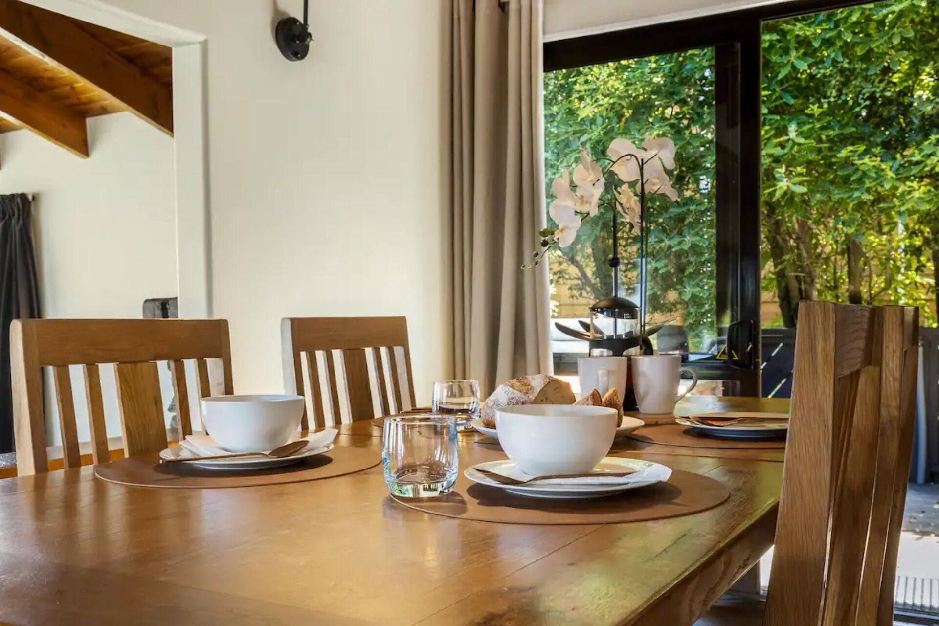 Une table à manger dressée avec des bols et des assiettes blancs, un verre d’eau, des chaises et une fenêtre donnant sur un paysage verdoyant.