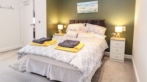 A neatly made bed with a patterned duvet, two bedside tables with lamps, a painting on the wall, and a door with a sign. - Kingfisher Lodge (Great Yarmouth)