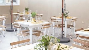 An outdoor dining area with white tables and chairs, beige umbrellas, and potted plants. - Tabernabè (Tortoli)