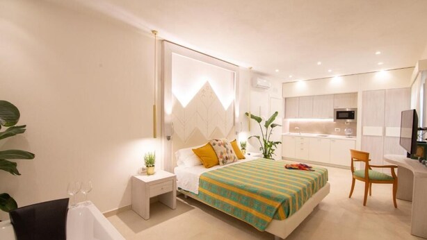 A modern bedroom with a large bed, bedside tables, a desk, and a kitchen area in the background. - Tabernabè (Tortoli)
