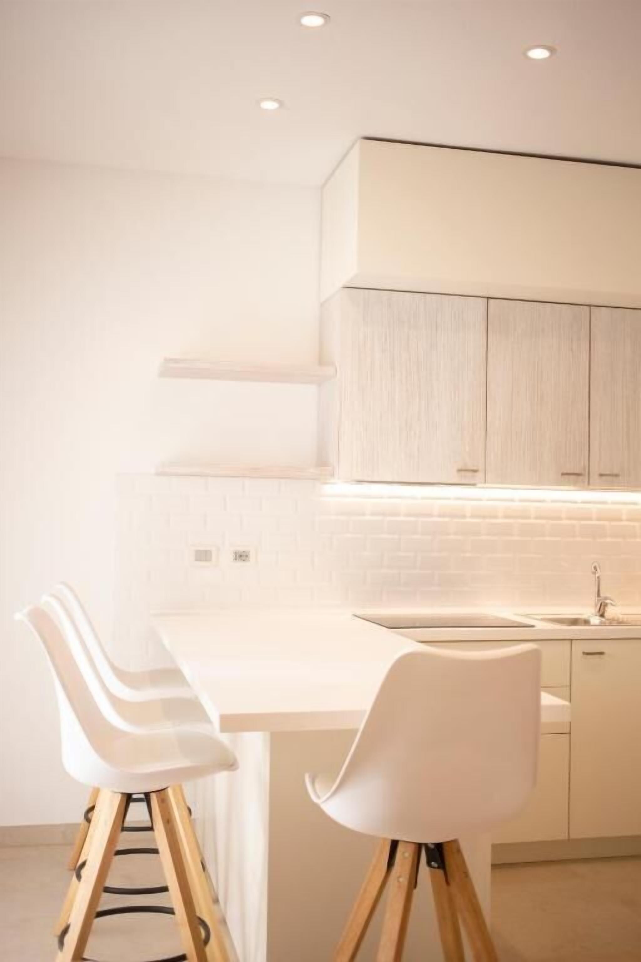 A modern kitchen with white cabinetry, a white countertop, and two white chairs with wooden legs.