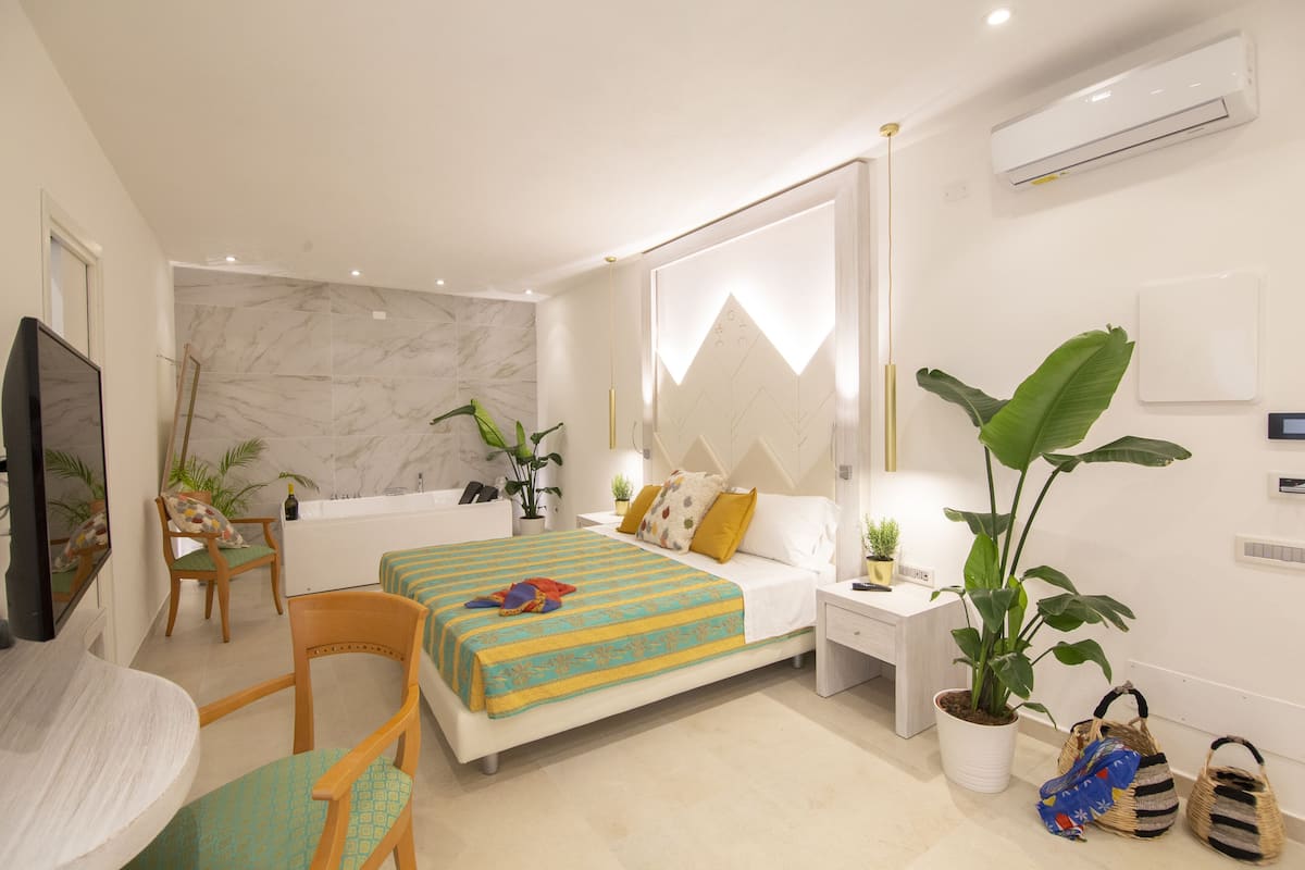 A modern bedroom with a bed, bedside tables, a desk, chairs, and potted plants.