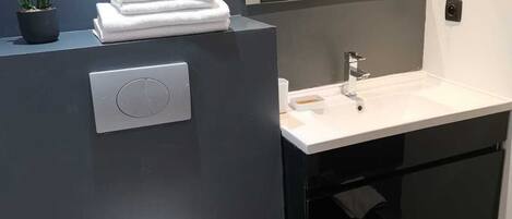 Un baño moderno con pared oscura, inodoro blanco, lavamanos con espejo y una pequeña planta en maceta sobre el mostrador.