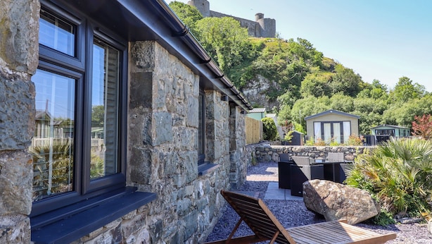 A stone building with large windows, a wooden lounge chair, and a view of a castle on a hill. - Tan Llech (Harlech)
