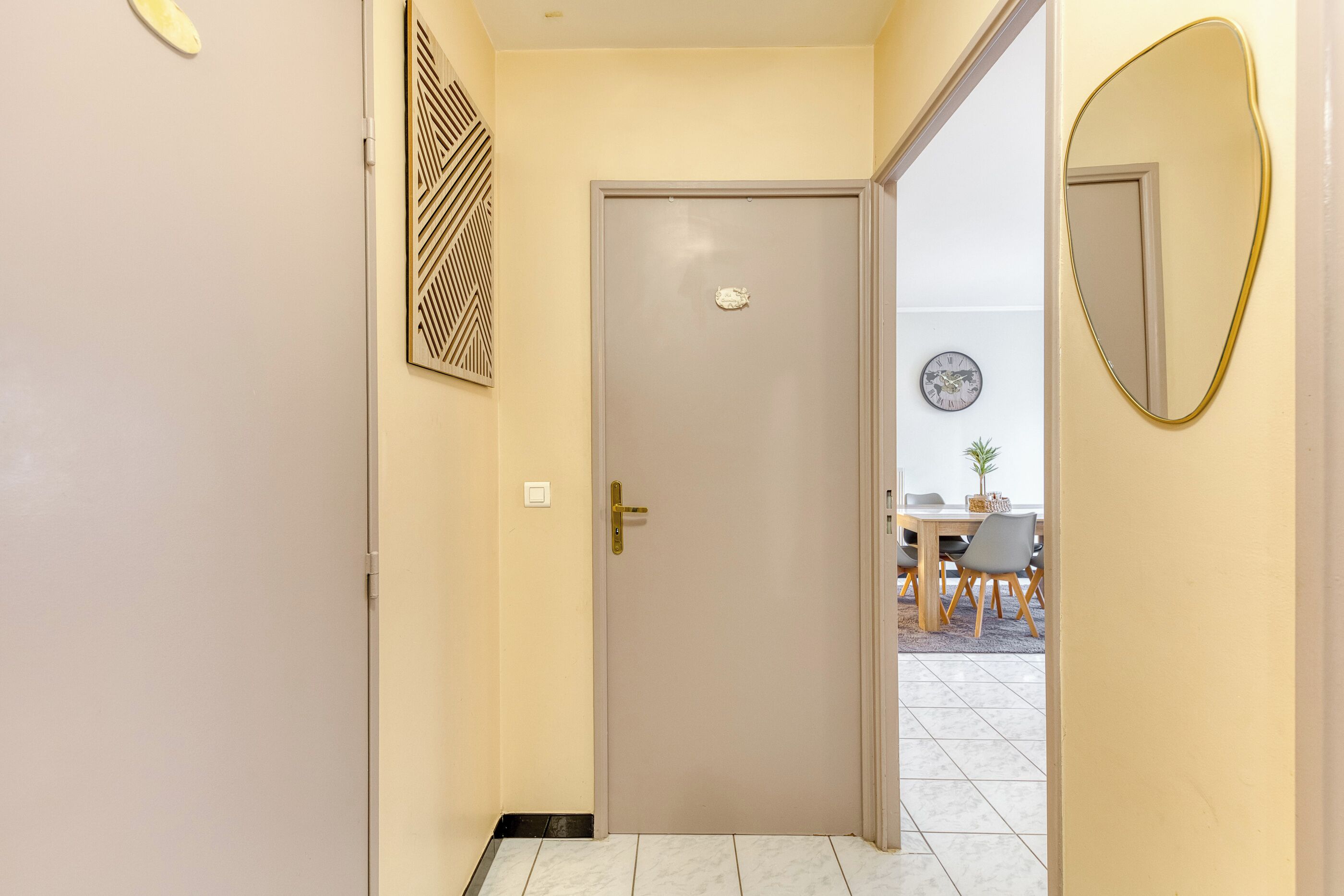 Un pasillo con una puerta cerrada, un reloj en la pared y un comedor visible a través de la puerta.