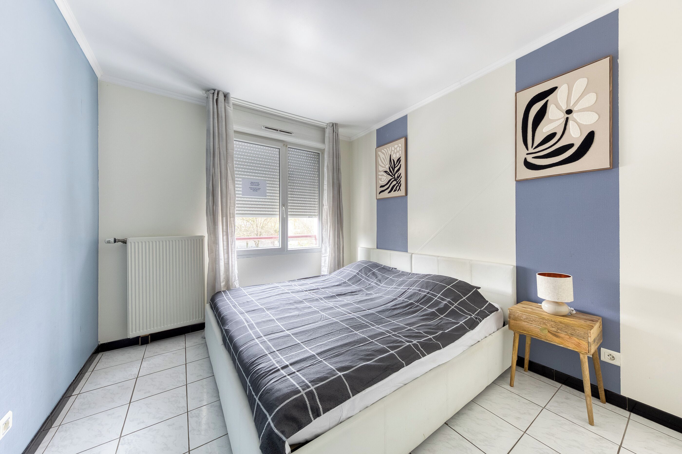 Une chambre à coucher comprenant un lit, une table de chevet, un radiateur, une fenêtre avec des rideaux et un mur orné d’un tableau encadré.