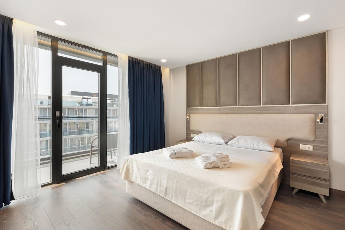 Quarto de hotel com uma cama grande, mesa de cabeceira, porta de vidro deslizante para uma varanda e vista da cidade.