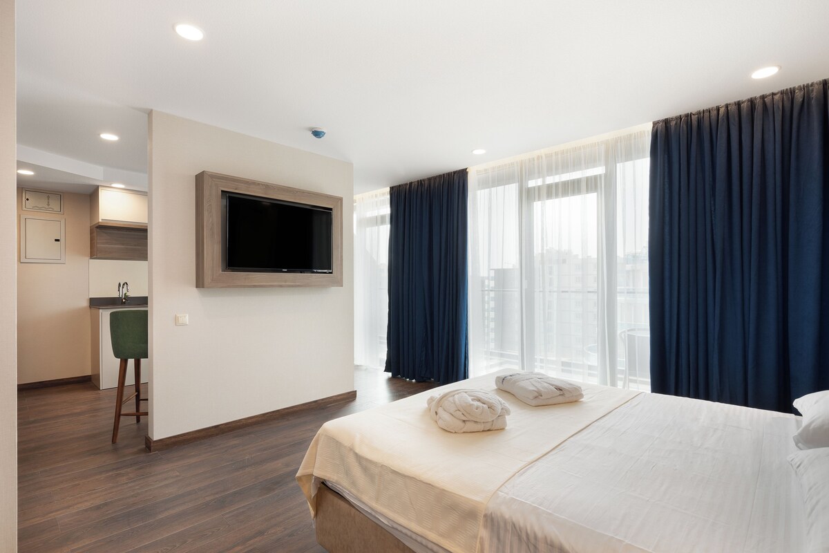 A hotel room with a bed, a TV mounted on the wall, a kitchen area, and large windows with curtains.