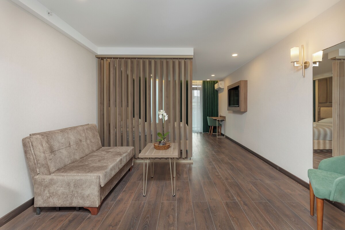 A modern living room with a sofa, a small table with a plant, a TV mounted on the wall, and a wooden floor.