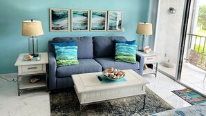 A living room with a blue sofa, two side tables with lamps, a coffee table with a bowl of seashells, and a large window with a view of greenery. - Beach Front Condo with Ocean View (Freeport)