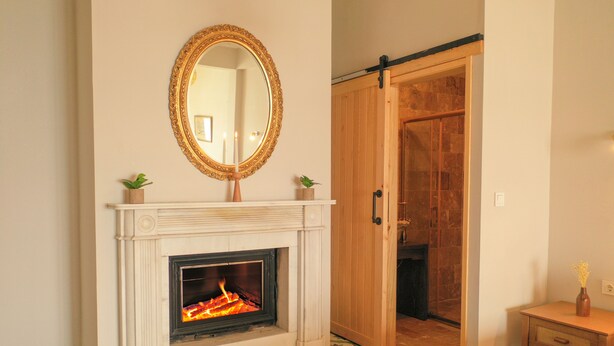 A room with a fireplace, a mirror, a wooden door, and a small table with a vase. - Ank'Ada Ayanikola (Adalar)