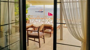 Vue depuis une pièce dont la fenêtre donne sur un balcon. Le balcon est aménagé avec des meubles en bois et une table, et on aperçoit un bateau arborant le drapeau turc sur l’eau.