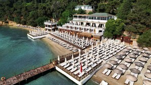 Un hôtel situé sur la côte, avec plusieurs étages, une jetée qui s’avance dans la mer, ainsi que de nombreux transats et parasols.
