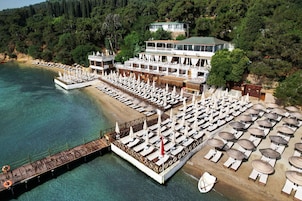 Un hotel costero de varios niveles, con un muelle que se adentra en el mar y numerosas tumbonas y sombrillas.