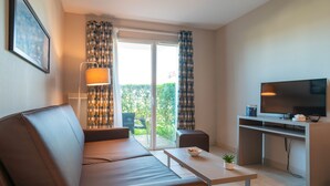 A modern living room with a sofa, a coffee table, a TV, and a balcony with curtains. - Les Thermes 508 - Appt avec piscine partagée (Concarneau)