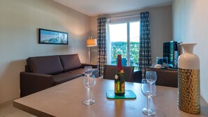 A modern living room with a sofa, a dining table with wine glasses and a bottle, and a large window with curtains. - Les Thermes 508 - Appt avec piscine partagée (Concarneau)